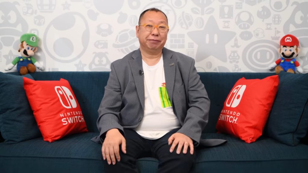 A man in a gray blazer sits on a blue couch with red Nintendo Switch pillows and Mario plush toys on the back wall behind him.