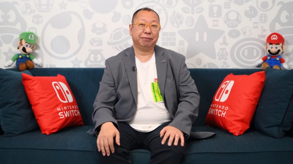 A man in a gray blazer sits on a blue couch with red Nintendo Switch pillows and Mario plush toys on the back wall behind him.