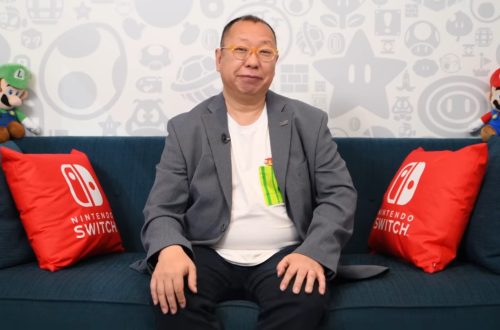 A man in a gray blazer sits on a blue couch with red Nintendo Switch pillows and Mario plush toys on the back wall behind him.